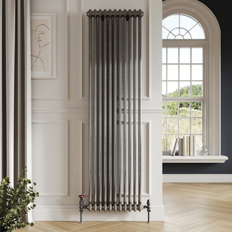 A vertical raw metal column radiator mounted against a white square panelled wall on light wood herringbone floor. A window can be seen in the background showing a bright countryside view.