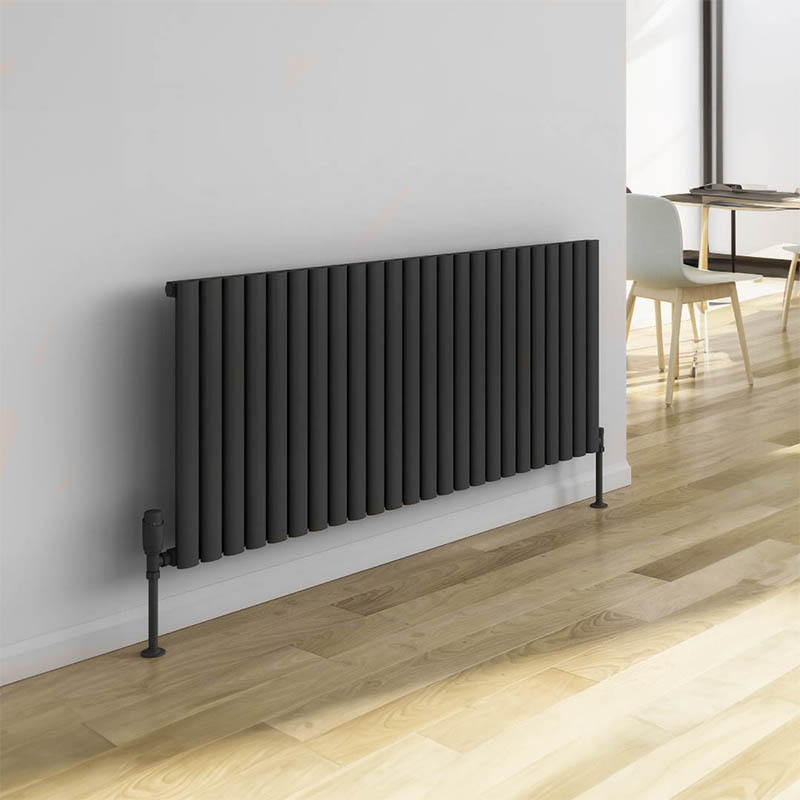A horizontal dark grey radiator featuring 24 slim, vertical columns. Mounted on warm wooden floorboards against a plain white wall.