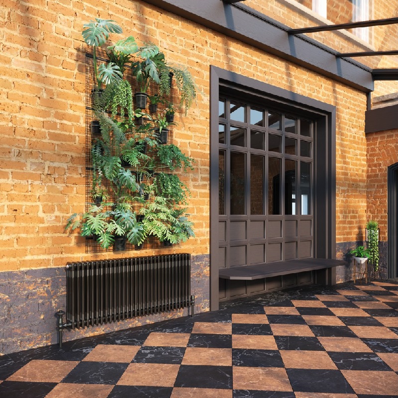 A black nickel column radiator mounted on a bare brick wall. The floor is terracota and black checkerboard tiles and their is a painted grey stripe on the bottom half of the brick wall.