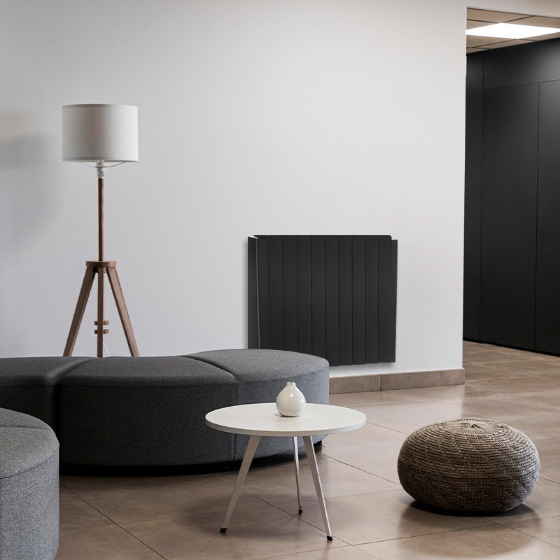 Pictured is a 1.8kW dark anthracite 10 panel radiator mounted on a plain light grey wall. It is situated with an abstract living room featuring a tall wooden-stand lamp behind a curved, low black sofa and white 3-legged roudn table.