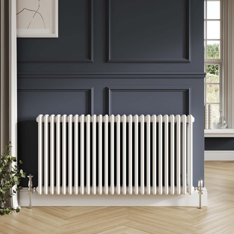 A white column radiator mounted against a navy square panelled wall on light wood herringbone floor. A window can be seen in the background showing a bright countryside view.